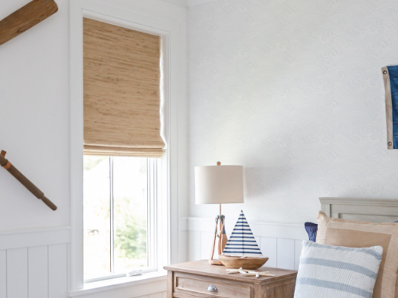 The image shows a bedroom interior with a white walls and ceiling, a large window with white blinds partially drawn, a wooden cross hanging on the wall above the window, a blue flag or banner with a white border to its right, a wicker basket with handles on top of a small table, and a bed with a simple headboard that appears to be made from wood. The room has a cozy feel with natural light coming through the window.