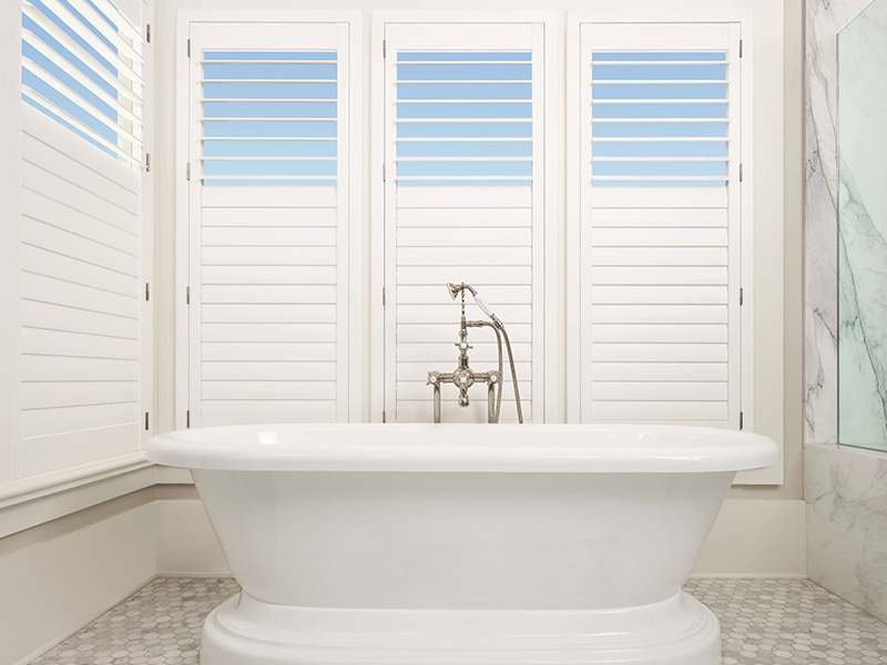 An image of a modern bathroom with a large white bathtub, a window with horizontal blinds, and a marble countertop.